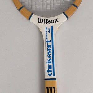 Puede incluir: Raqueta de tenis Wilson Chris Evert American Star vintage. Cuenta con un marco de madera con un mango blanco y azul. La raqueta tiene un agarre negro y cuerdas plateadas. El logotipo de Wilson es visible.