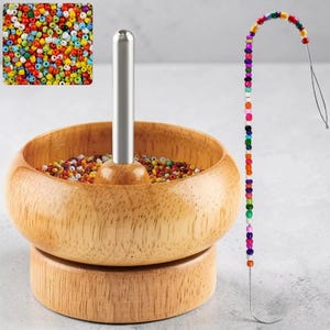 Op de afbeelding: Een houten kralenspin met een roestvrijstalen naald en een verscheidenheid aan kleurrijke kralen. De spinner is gevuld met kleine, veelkleurige kralen. Een rij kralen is op een naald geregen, wat het rijgproces laat zien.