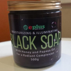 Könnte beinhalten: Ein schwarzes Glas "Black Soap" mit goldenem Deckel. Das Etikett enthält den Text "Moisturizing & Illuminating" und "With Honey and Papaya Oil For a Radiant Complexion". Das Glas enthält 500g Produkt.