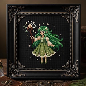 May include: A framed cross-stitch artwork featuring a green-haired fairy holding a staff with glowing orbs. The fairy wears a green dress and stands against a black background. The frame is black with ornate corner details. A small glass bottle and a leather-bound book are in the foreground.