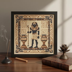 May include: Framed cross-stitch artwork featuring an Egyptian pharaoh figure in black, tan, and gold. The artwork includes hieroglyphs and decorative borders, presented in a black frame. Two decorative urns flank the pharaoh. The artwork is displayed on a wooden surface.