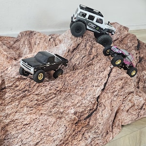 May include: Four remote-controlled off-road vehicles on a textured brown rock-patterned surface. The collection includes a blue SUV, a black pickup truck with gold wheels, a white SUV with black accents, and a small pink and black crawler.