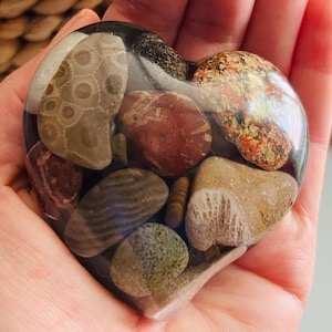 May include: A heart-shaped resin paperweight containing a collection of smooth, multi-colored river stones and fossils. The stones feature various textures and patterns, including circular fossil imprints, speckled red, and grey striped designs, all encased in a clear, polished resin finish.