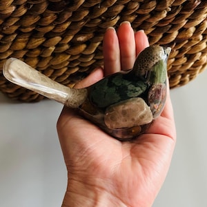 May include: A bird-shaped decorative object crafted from clear resin, containing various smooth, multi-colored natural stones and pebbles embedded within its body, featuring a long, light-colored, textured tail section.