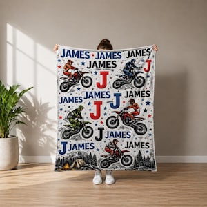 May include: A white blanket featuring a motocross theme with the name "JAMES" repeated. The design includes illustrations of riders on motorcycles, the letter "J", stars, and a tent scene. The color scheme includes red, blue, and black.