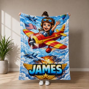 Puede incluir: Una manta vibrante con una ilustración de dibujos animados de un niño en un avión rojo y amarillo, volando por un cielo azul con nubes, aviones y un globo aerostático. La manta tiene el nombre "JAMES" en letras grandes.