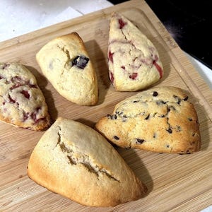May include: Five freshly baked scones on a wooden cutting board. The scones vary in color and texture, with some featuring chocolate chips and others with visible berries. The baked goods are golden brown and triangular in shape.