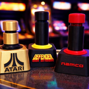 May include: Three arcade joystick controllers with different designs. One is gold with the Atari logo, another is black with "DEFENDER" in red and yellow, and the third is black with "namco" in red.