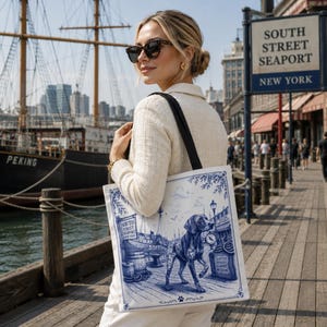 May include: A white canvas tote bag featuring a blue ink-style illustration of a dog standing on a dock. The graphic includes nautical elements, barrels, a street lamp, and text reading South Street Seaport and South Street Seaport Museum.