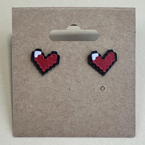 May include: A pair of pixelated heart earrings. Each earring features a red heart shape with a black outline and a white accent. The earrings are displayed on a brown cardboard earring card.