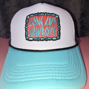May include: A trucker hat with a light blue brim and a white front panel. The front features a rectangular patch with a red background, a teal border, and the embroidered teal text "LONG LIVE COWBOYS". A black cord trim separates the brim and crown.