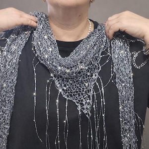 May include: A shimmering silver scarf with a delicate, open weave design. The scarf features sparkling embellishments and long, dangling strands. The scarf is draped over a black top, creating a contrast of textures and colors.
