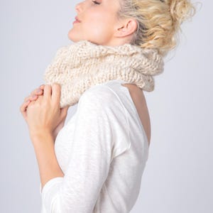 Könnte beinhalten: Ein cremefarbener, grob gestrickter Rundschal liegt um den Hals einer Person, die ein weißes Langarmshirt trägt. Der Schal wirkt weich und warm, ideal für kühleres Wetter. Die Person hat die Augen geschlossen.