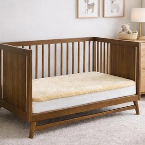 May include: A wooden crib with a light-colored mattress and a fluffy, cream-colored cover. The crib has vertical slats and a low profile. The room has framed animal art and a small lamp.