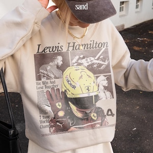 Peut inclure: Sweat-shirt beige avec un imprimé graphique de Lewis Hamilton, pilote de Formule 1. Le design comprend des images de Hamilton, son casque et le texte "Lewis Hamilton", "We Win And Lose Together", et "7 Time World Champion".