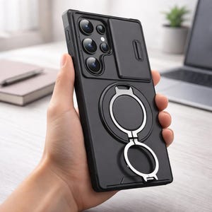 May include: Black phone case with a sliding camera cover and a silver ring holder. The case has a rectangular shape and is designed to protect the phone's camera lenses. The ring holder can be used as a stand or for a secure grip.