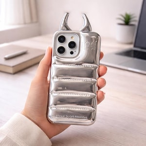 Op de afbeelding: Zilveren telefoonhoesje met een uniek ontwerp, met gebogen hoorns en een gewatteerde textuur. De hoes heeft een camera-uitsparing en de tekst "Urban Sophistication". De hoes wordt in een hand gehouden.