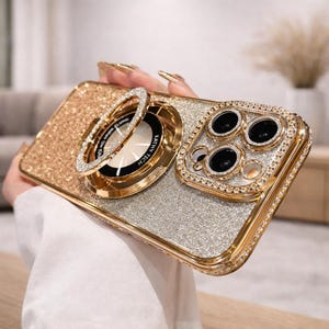 Könnte beinhalten: Eine gold- und silberfarbene Glitzer-Handyhülle mit Ringhalter und Kameraschutz. Die Hülle hat einen goldenen Rahmen und ist mit Strasssteinen besetzt. Der Ringhalter ist goldfarben und hat ein diamantartiges Design.