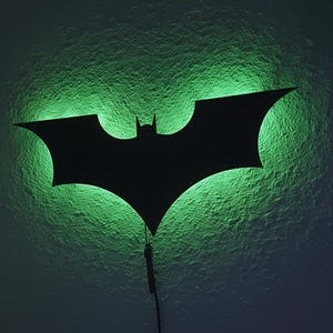 Puede incluir: Luz de pared con el logotipo de Batman negro con brillo de neón verde. La silueta está montada en una pared texturizada, con un cable de alimentación que se extiende hacia abajo. Un artículo decorativo para un dormitorio o sala de juegos.