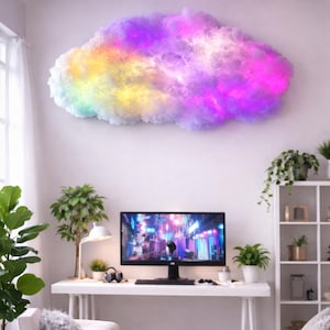 Puede incluir: Una lámpara de pared con forma de nube con una vibrante exhibición de colores, incluyendo morado, amarillo y rosa. La nube está sobre un escritorio con un monitor de computadora, una lámpara de escritorio y plantas, creando un diseño interior moderno y colorido.