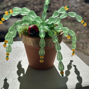 May include: A handcrafted beaded plant in a terracotta pot. The plant features light green, teardrop-shaped beads, accented with yellow and green beads. A small, red ladybug decoration sits in the pot. The pot has a decorative gold band.