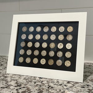 Può includere: Una cornice bianca contenente una collezione di monete d'argento, probabilmente da dieci centesimi, disposte a griglia su sfondo nero. Le monete sono presentate all'interno di una cornice di legno bianca, creando una presentazione ordinata. La cornice è rettangolare e poggia su una superficie maculata.