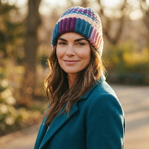 Op de afbeelding: Een vrouw draagt een kleurrijke, gehaakte beanie met tinten paars, blauw en roze. Ze draagt ook een teal jas. De achtergrond is wazig, wat wijst op een buitenomgeving, mogelijk een park of tuin.