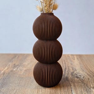 May include: A brown, stacked vase with a ribbed design, holding dried beige flowers. The vase has a unique, three-sectioned shape. The arrangement sits on a wooden surface, creating a natural, earthy aesthetic. The vase is approximately 12 inches tall.