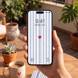 Pu&ograve; includere: Uno smartphone mostra uno sfondo a righe blu chiaro e bianco con un cuore rosso. Lo schermo indica le 9:41, luned&igrave; 6 gennaio. Il telefono &egrave; tenuto in mano, con una tazza di caff&egrave; e una piccola pianta in vaso visibili.