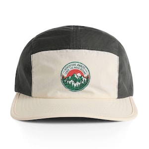 May include: A five-panel cap with a dark grey crown, a cream-coloured bill, and a cream-coloured front panel. The front panel features a circular patch with a mountain and forest design and the text "ADVENTURE AWAITS LEAVE THE NOISE BEHIND".