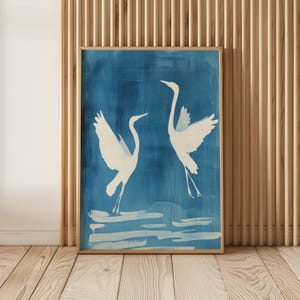 Puede incluir: Una impresión artística enmarcada con dos garzas blancas sobre un fondo azul. Las aves se representan con las alas extendidas, sugiriendo movimiento. La obra de arte está enmarcada en un tono de madera clara, añadiendo un toque natural. El estilo recuerda a una acuarela.