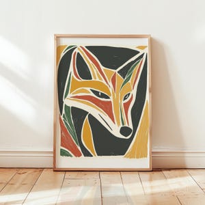 May include: A framed art print featuring a stylized fox illustration in a block print style. The fox is rendered with bold, geometric shapes in shades of orange, yellow, and dark green against a deep charcoal background, all encased in a light wood frame.