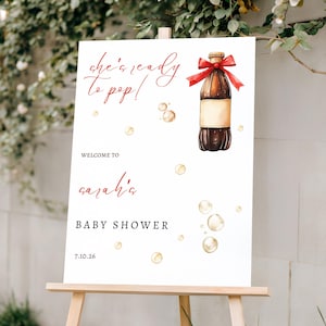 May include: A white baby shower sign on a wooden easel. The sign features the text "she's ready to pop!" and "Welcome to Sarah's Baby Shower 7.10.26". A watercolor illustration of a soda bottle with a red bow is on the sign.