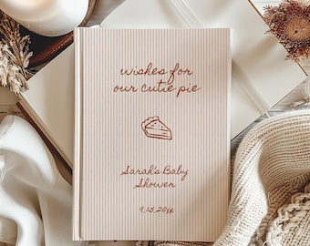 May include: A rectangular guest book with a light beige and white pinstripe cover. The text reads: wishes for our cutie pie, Sarah's Baby Shower, and 9.15.2024. A brown line drawing of a slice of pie is centered between the text lines.
