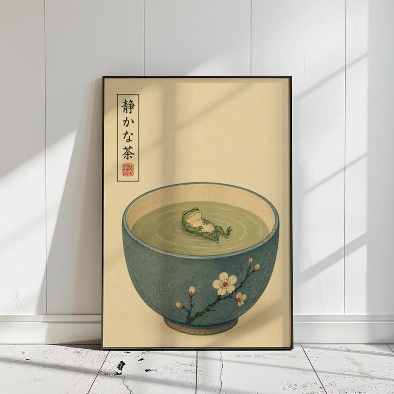 A black-framed vertical print of a frog in a tea bowl hangs on a white paneled wall above a tiled floor.