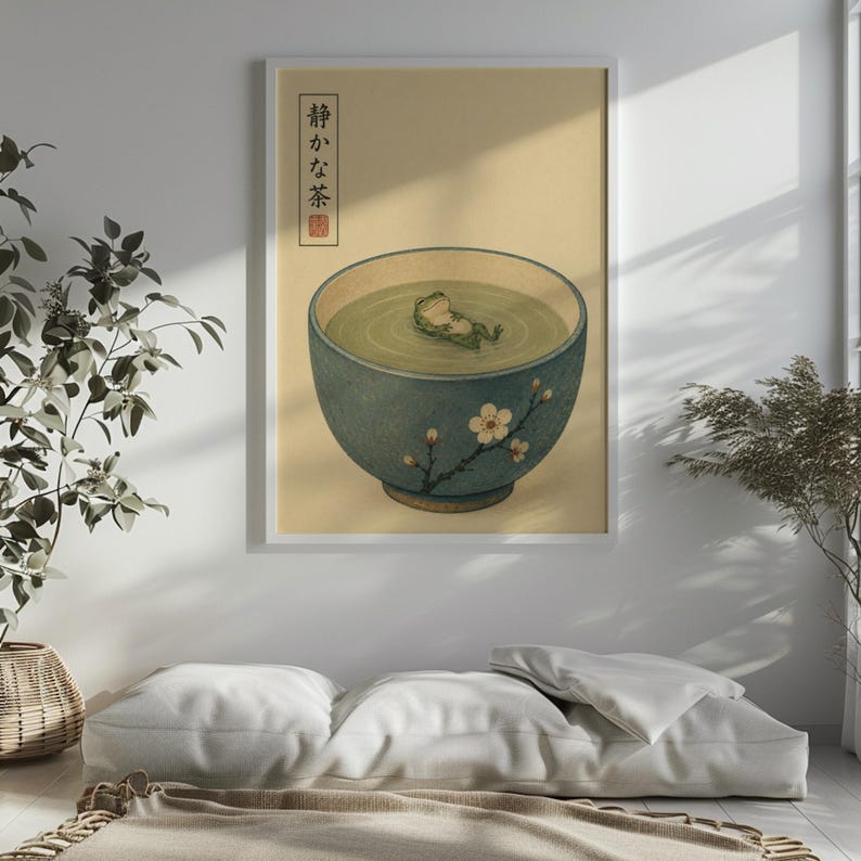 A framed print of a frog in a bowl hangs on a white wall above floor cushions and next to indoor plants.