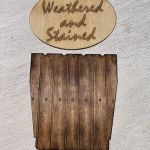 Peut inclure: Panneau en bois avec les mots "Weathered and Stained" grav&eacute;s sur un ovale. En dessous, une pi&egrave;ce de bois ressemblant &agrave; un c&ocirc;t&eacute; de tonneau, avec des lignes verticales et de petits d&eacute;tails circulaires. Le bois est teint&eacute; brun fonc&eacute;.