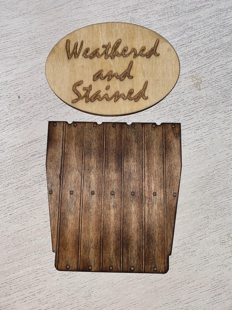 Peut inclure: Panneau en bois avec les mots "Weathered and Stained" grav&eacute;s sur un ovale. En dessous, une pi&egrave;ce de bois ressemblant &agrave; un c&ocirc;t&eacute; de tonneau, avec des lignes verticales et de petits d&eacute;tails circulaires. Le bois est teint&eacute; brun fonc&eacute;.