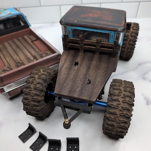 May include: A remote-controlled truck with a weathered blue and brown cab, a dark wood-grain flatbed, and large knobby tires. A separate brown truck bed with wood-grain detailing sits nearby, accompanied by four small black L-shaped metal brackets.