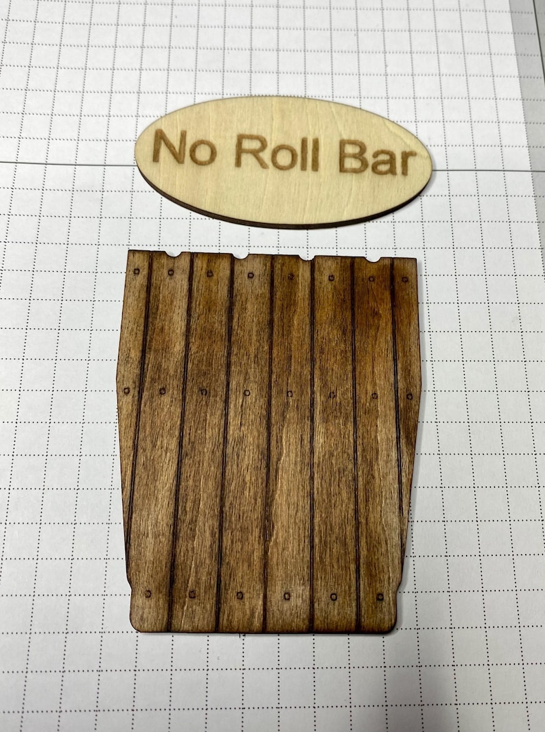 May include: A light-colored oval wooden plaque with the text No Roll Bar laser-engraved in brown, positioned above a dark-stained wooden piece featuring vertical slats and small circular details resembling nail heads in a grid pattern.