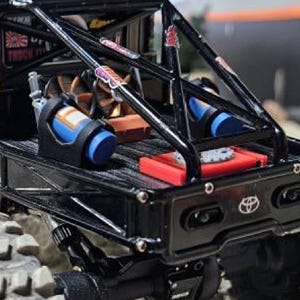 May include: A black remote-controlled truck bed featuring a roll cage, a blue nitrous oxide canister, a red battery pack, and a Toyota emblem on the tailgate. The truck bed includes a cooling fan and various stickers attached to the metal frame.