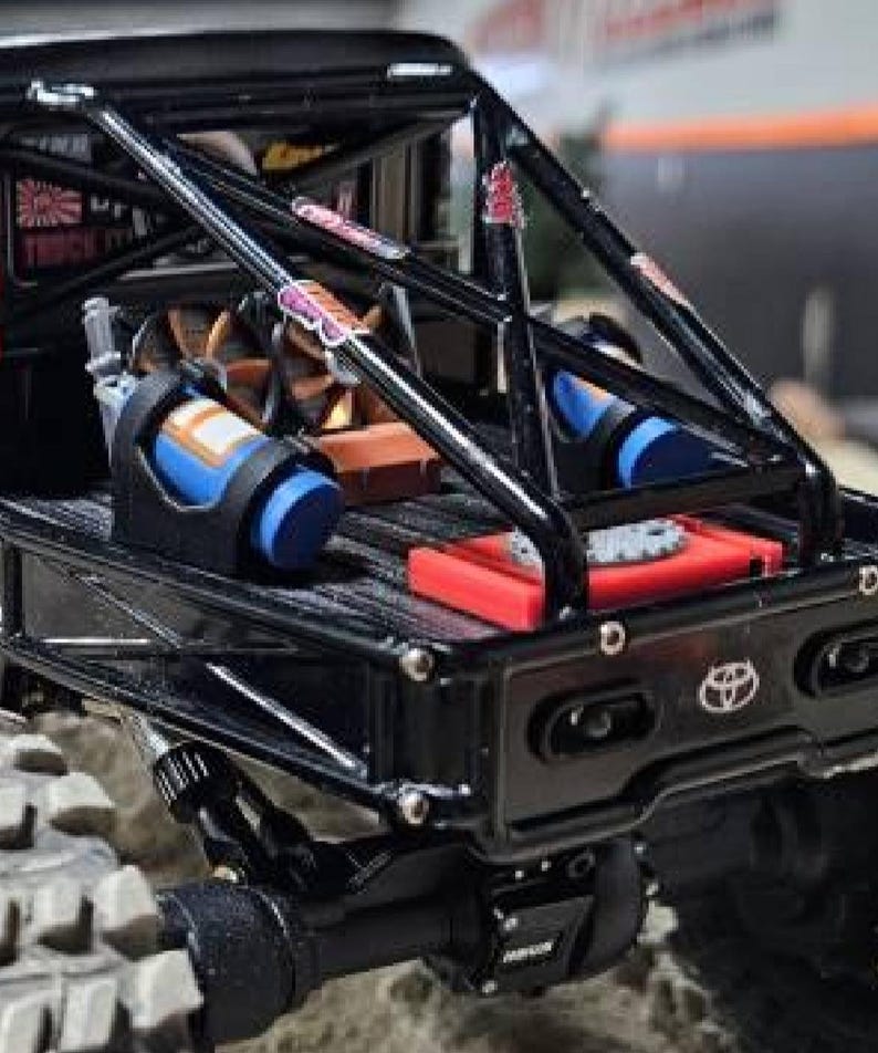 May include: A black remote-controlled truck bed featuring a roll cage, a blue nitrous oxide canister, a red battery pack, and a Toyota emblem on the tailgate. The truck bed includes a cooling fan and various stickers attached to the metal frame.