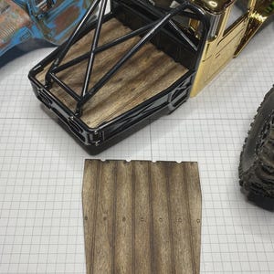 May include: A scale model truck bed featuring a wood-grain floor and a black metal roll cage, accompanied by a separate, matching rectangular wood-grain panel with small circular rivet details along the edges for assembly.