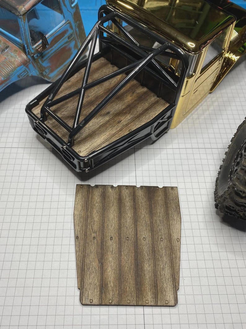 May include: A scale model truck bed featuring a wood-grain floor and a black metal roll cage, accompanied by a separate, matching rectangular wood-grain panel with small circular rivet details along the edges for assembly.