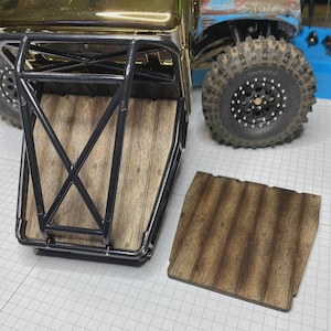 May include: A remote-controlled truck bed featuring a black metal roll cage frame and a wood-grain textured floor panel. A matching removable wood-grain panel is placed beside the truck, which has large black off-road tires and a gold hood.