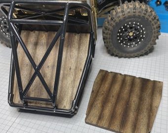 May include: A remote-controlled truck bed featuring a black metal roll cage frame and a wood-grain textured floor panel. A matching removable wood-grain panel is placed beside the truck, which has large black off-road tires and a gold hood.