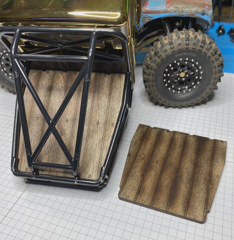 May include: A remote-controlled truck bed featuring a black metal roll cage frame and a wood-grain textured floor panel. A matching removable wood-grain panel is placed beside the truck, which has large black off-road tires and a gold hood.