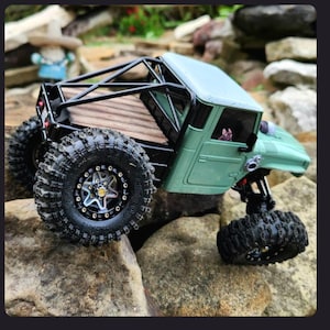 May include: A sage green remote-controlled crawler truck featuring a black roll cage, a wooden truck bed, and large, knobby black tires with silver rims. The vehicle has a detailed cabin interior and a small figure visible through the window.