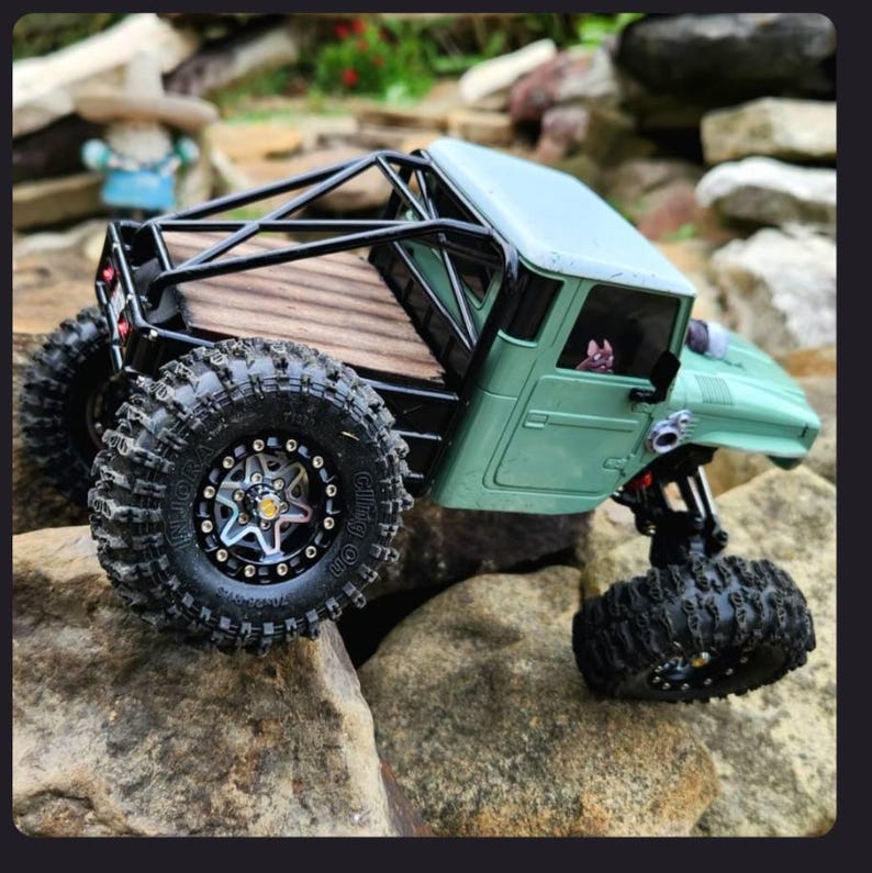 May include: A sage green remote-controlled crawler truck featuring a black roll cage, a wooden truck bed, and large, knobby black tires with silver rims. The vehicle has a detailed cabin interior and a small figure visible through the window.