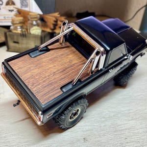 May include: A black toy pickup truck with a wooden bed and chrome roll bar. The truck has large, off-road tires and a detailed design. The bed of the truck has a wood grain pattern.